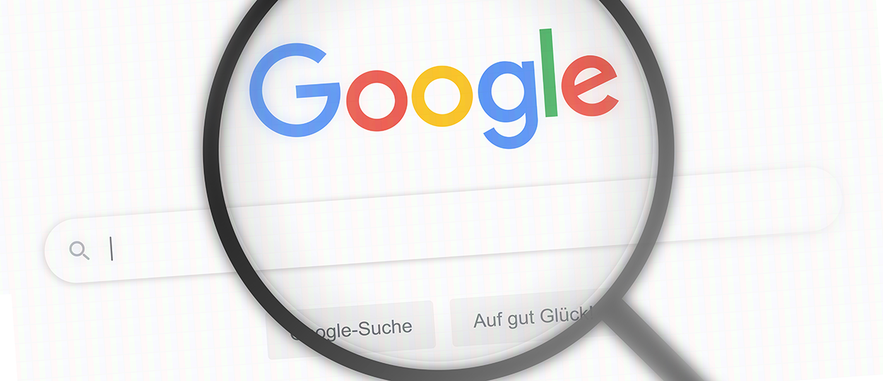 Bildbeschreibung: Das Bild zeigt eine Nahaufnahme des Google-Logos auf einer Suchseite, vergrößert durch eine Lupe, was den Fokus auf die Google-Suche legt.