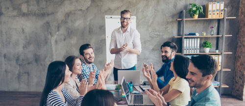 Bildbeschreibung: Das Foto zeigt eine Gruppe von Menschen, die in einem modernen Büro an einem Tisch sitzen und einem lächelnden Mann applaudieren, der vor ihnen steht und offenbar eine Präsentation oder ein Meeting leitet.