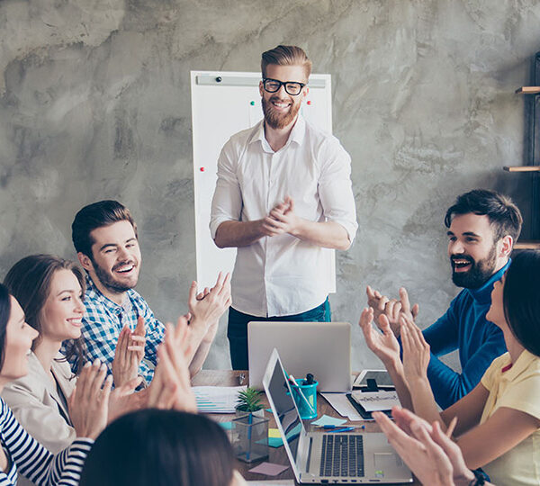 Bildbeschreibung: Das Foto zeigt eine Gruppe von Menschen, die in einem modernen Büro an einem Tisch sitzen und einem lächelnden Mann applaudieren, der vor ihnen steht und offenbar eine Präsentation oder ein Meeting leitet.